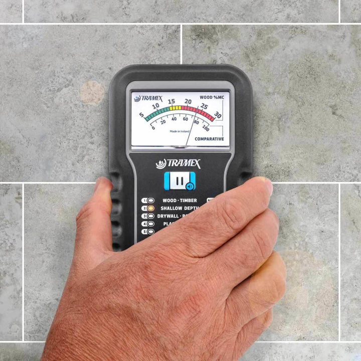 A hand holding the Tramex ME5 moisture meter over gray tiled flooring