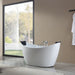 Modern white freestanding bathtub with curved design, positioned near large floor-to-ceiling window overlooking trees and garden.