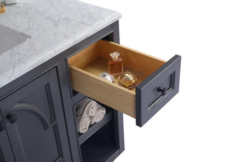 Bathroom vanity with open drawer holding perfume and creams, gray cabinet, marble countertop, and towels on shelf.