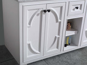 Close-up of a white vanity with decorative cabinet doors, black knobs, a drawer, and an open shelf with toiletries.
