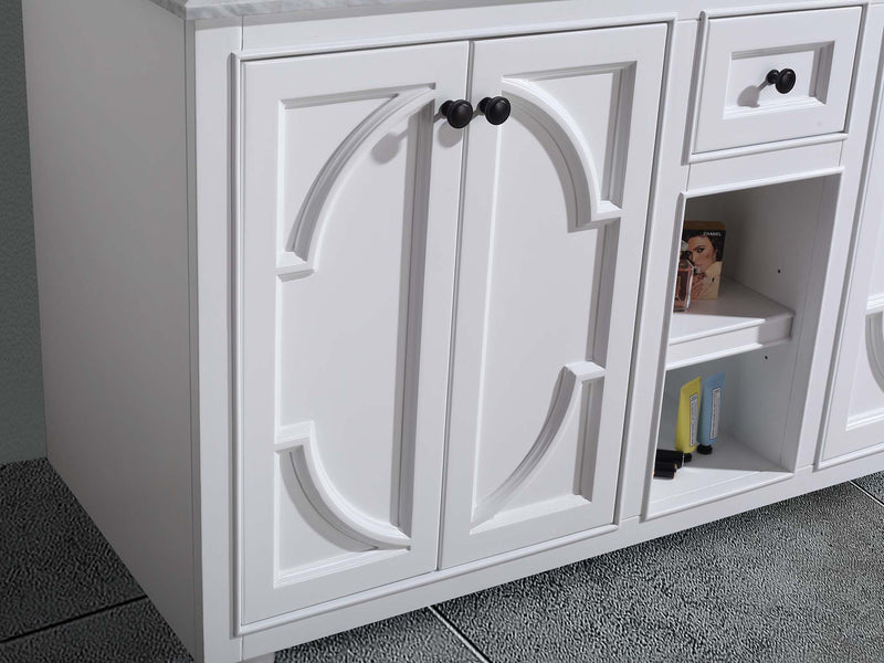Close-up of a white vanity with decorative cabinet doors, black knobs, a drawer, and an open shelf with toiletries.
