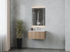 Modern bathroom vanity with wooden cabinet, white countertop, illuminated mirror, potted plant, and gray tile walls.
