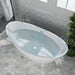 Modern white freestanding oval bathtub with blue-tinted water, set on hexagonal marble tiles with gray subway tile wall and green plant accent.