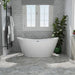 Modern white freestanding bathtub against gray brick wall, with French windows, towel rack, and potted plants creating a serene bathroom setting.