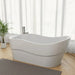 Modern white freestanding bathtub with curved design, placed on wooden floor with gray mat and green potted plant, soft curtains in background
