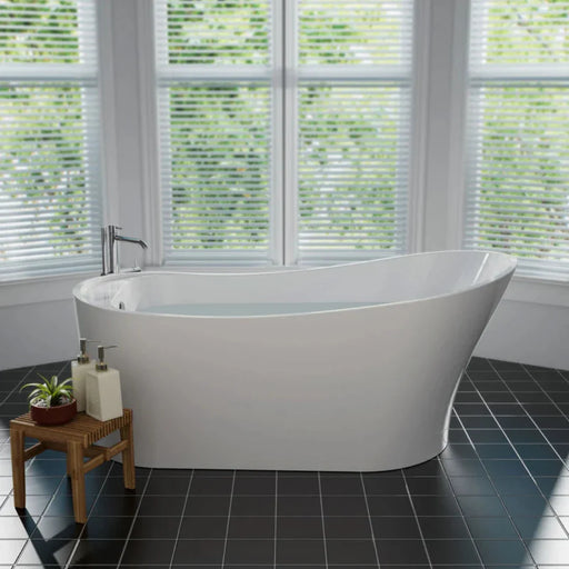 Modern white freestanding bathtub beside window with blinds, positioned on dark tiled floor with wooden stool and bathroom accessories.