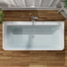 Modern white rectangular bathtub with chrome faucet, viewed from above on wooden floor with hints of green plant leaves.