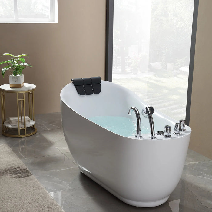 Modern white freestanding bathtub with chrome fixtures, filled with water, and a black headrest, next to a window and side table with a green plant.