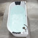 Modern white freestanding bathtub filled with light blue water, featuring black padded headrests and chrome fixtures on marble-like tiled floor.