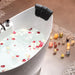 Tranquil bathtub scene with rose petals floating in water, surrounded by glowing candles on pebble-lined floor.