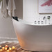 Modern white bathtub with lit candles on wooden floor, creating a relaxing spa-like bathroom atmosphere with white towels and a botanical art print.