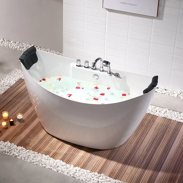 Modern white bathtub filled with water and flower petals, surrounded by wooden floor and white pebbles, with lit candles nearby.