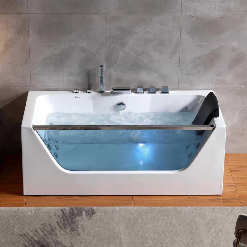 Modern white bathtub with glass side panel, filled with blue water and multiple chrome faucet controls, set against gray marble-tiled wall and wooden floor.