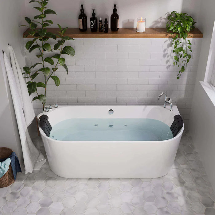 Modern white bathtub filled with blue water, surrounded by white subway tiles, hanging plants, and a wooden shelf with candle and dark bottles.