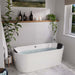 Modern white bathroom with freestanding tub, white subway tiles, wooden shelf with black bottles, and hanging green plants creating a serene spa-like atmosphere.