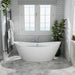 Modern white freestanding bathtub with gray tile walls, two windows, and green plants, creating a serene bathroom atmosphere.