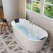 Modern freestanding white bathtub filled with water, positioned near large windows overlooking a serene lake and green landscape, with wooden stool and bath accessories nearby.