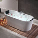 Modern white bathtub filled with water, scattered rose petals, surrounded by wooden decking and white pebbles, with lit candles nearby creating a relaxing spa-like atmosphere.