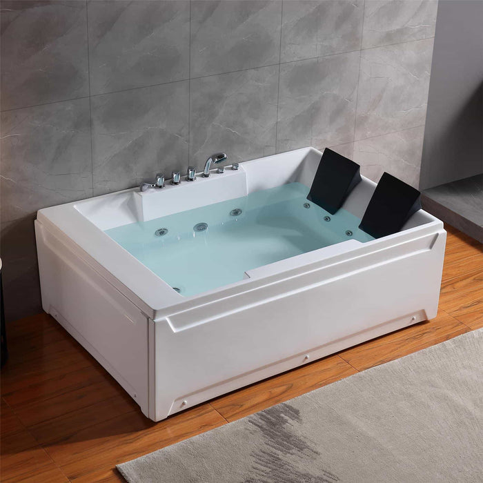 White two-person whirlpool bathtub filled with blue water, featuring chrome fixtures and black headrests, set on wooden flooring against gray tile wall.