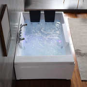 Large white whirlpool bathtub filled with swirling blue water, chrome fixtures, and two black headrests against modern bathroom tiles and wood flooring.