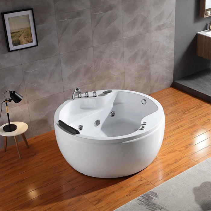 Modern white round bathtub with jets on wooden floor, gray tile wall, and small side table with desk lamp.