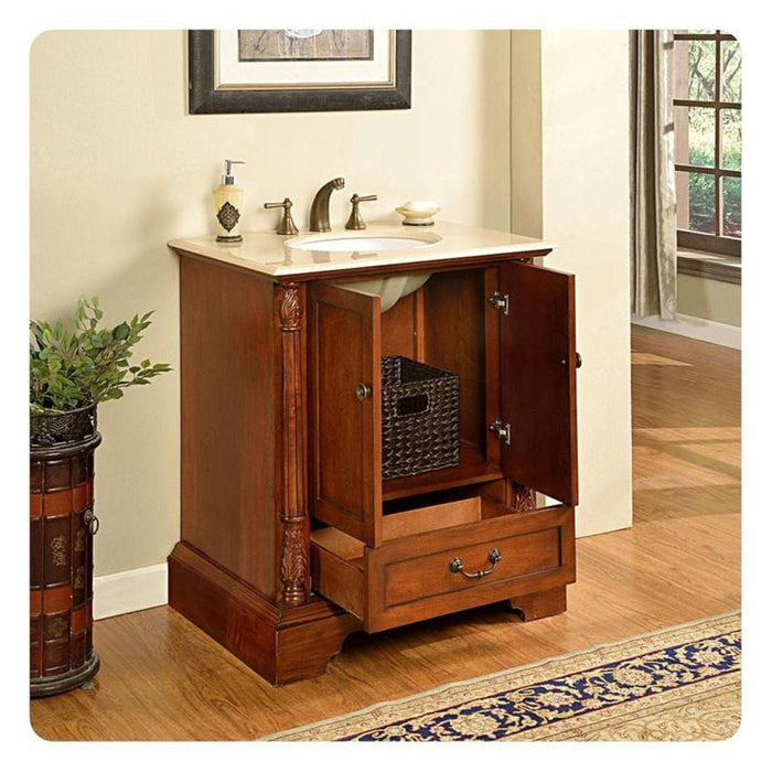 Wooden bathroom vanity with marble top, open cabinet revealing woven basket, ornate carved column, soap dispenser, and window in background.