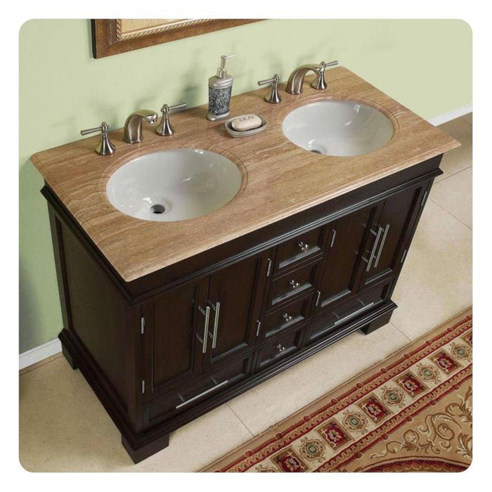 Dark wood bathroom vanity with travertine top, double white sinks, and chrome faucets, with decorative soap dispenser and rug.