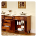 Rich wood bathroom vanity with double sinks, ornate drawers, floral artwork, and folded towels on open shelving.
