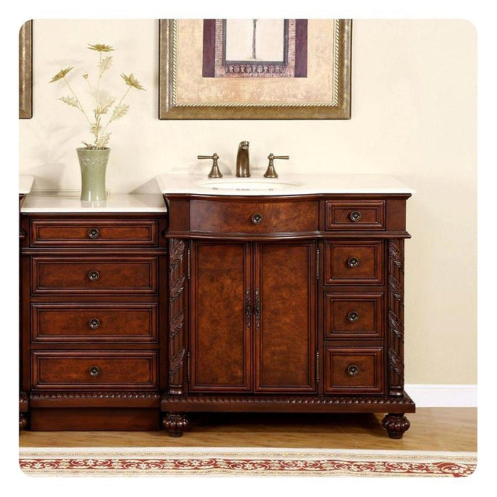 Rich mahogany bathroom vanity with detailed carved drawers, white marble top, and bronze sink fixtures, with a decorative flower vase and framed artwork.