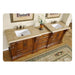 Ornate wooden bathroom vanity with two white sinks, travertine countertop, decorative vase with white flowers, and intricate area rug.