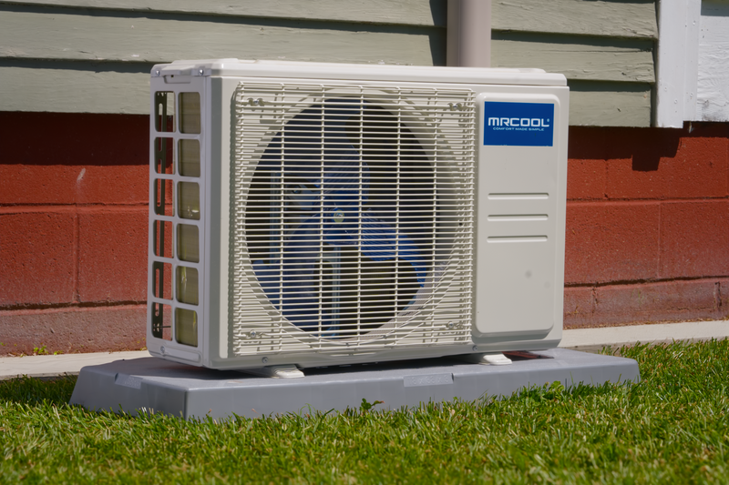 MRCOOL DIY Select condenser on a gray pad on the lawn beside a brick and painted-siding wall