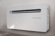 MRCOOL Monoblock mounted low on a painted interior wall above the baseboard, three-quarter view of the white cabinet and its slot air outlet