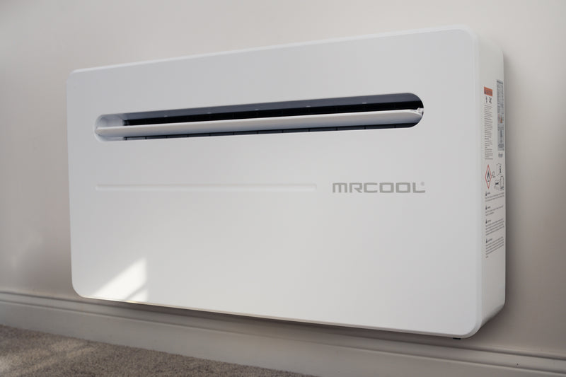 MRCOOL Monoblock mounted low on a painted interior wall above the baseboard, three-quarter view of the white cabinet and its slot air outlet